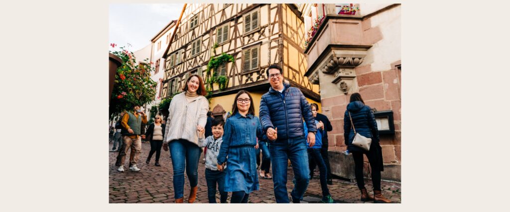 Family of four walking and holding hands through historic streets of Riquewihr, Alsace with timber-framed buildings in the background